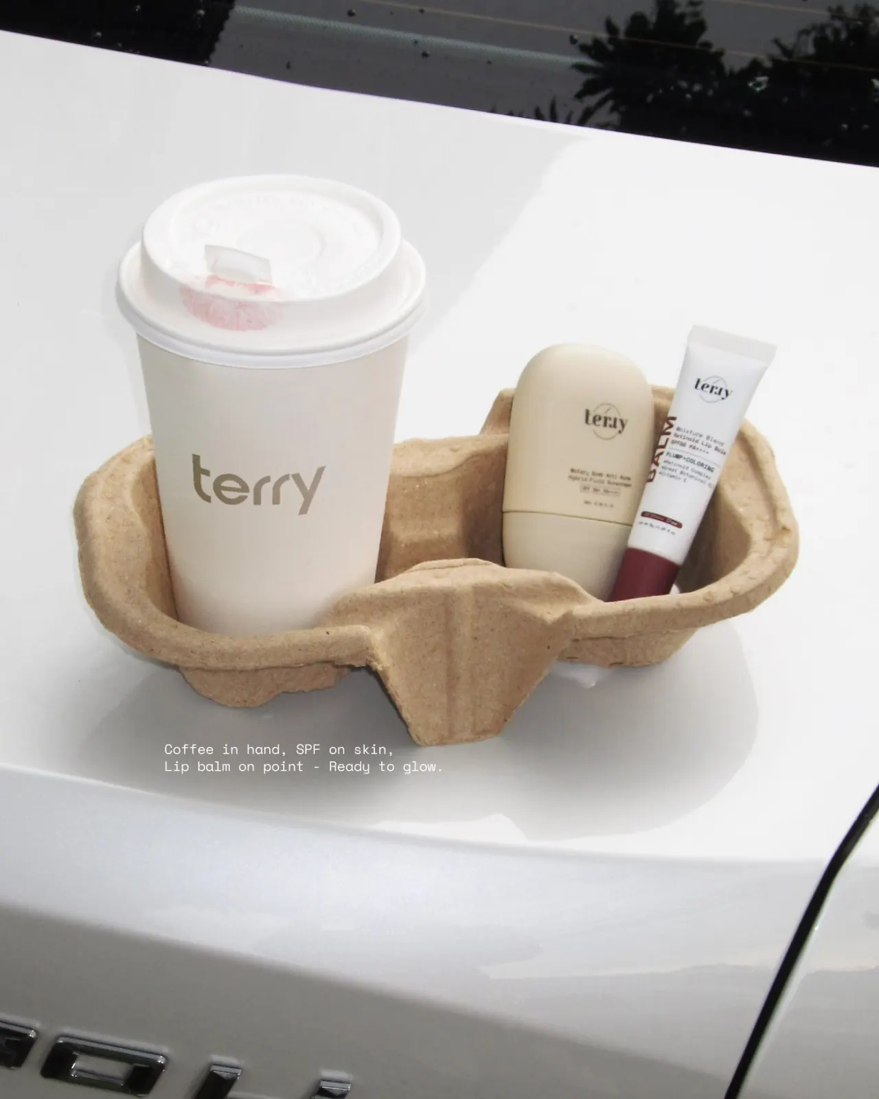 แก้วกาแฟโลโก้ terry วางในถาดกระดาษคู่กับกันแดดขนมปังและลิปบาล์มบนฝากระโปรงรถ