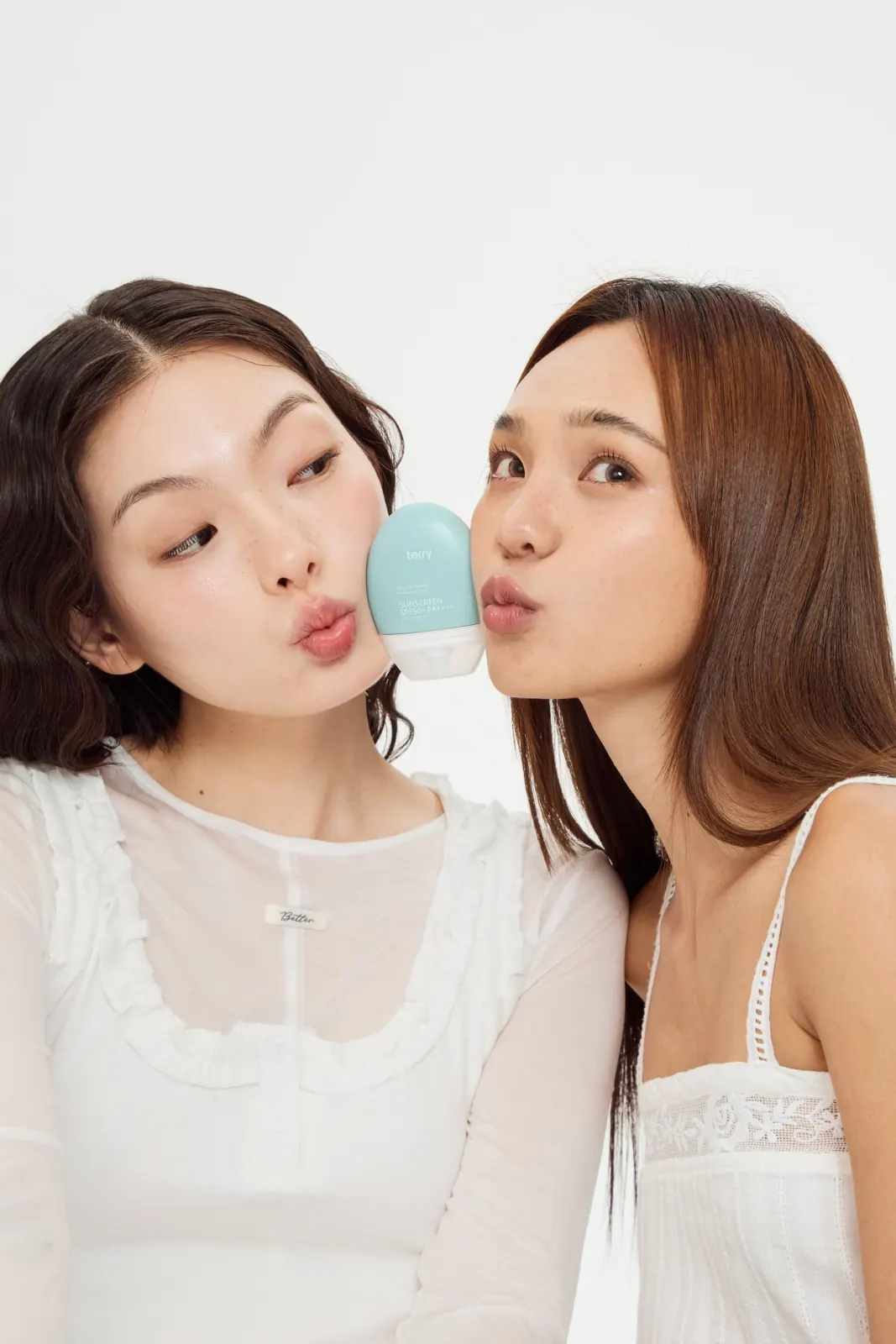 นางแบบสองคนหันเข้าหากัน ทำปากจู๊บ มีขวดกันแดดโทนอัพสีมิ้นต์คั่นอยู่ตรงกลาง