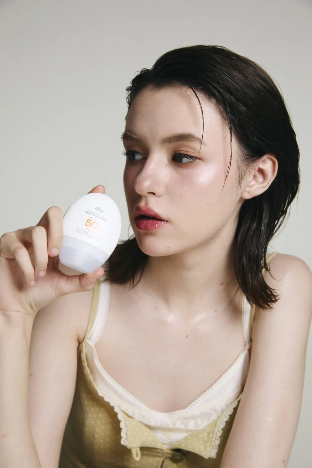 นางแบบถือขวด UV Defense ไว้ข้างแก้ม เห็นขนาดขวดเทียบกับใบหน้า